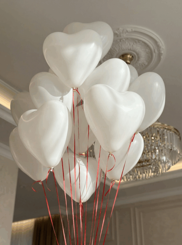 Белые латексные воздушные шары в форме сердца с красными свисающими лентами под потолком на фоне люстры.